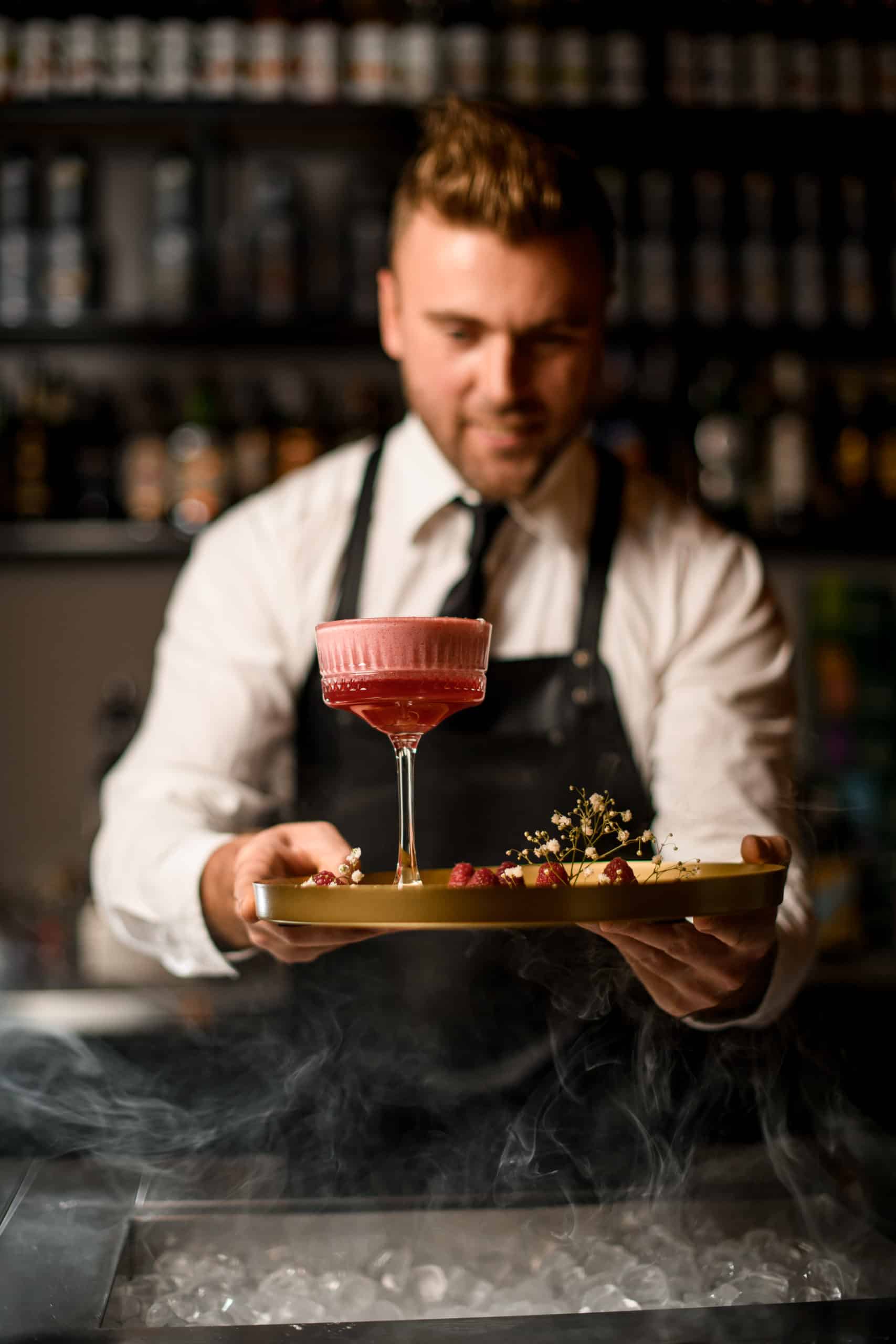 hand of barman holds round tray with raspberries and flowers and glass of pink foamy cocktail on it