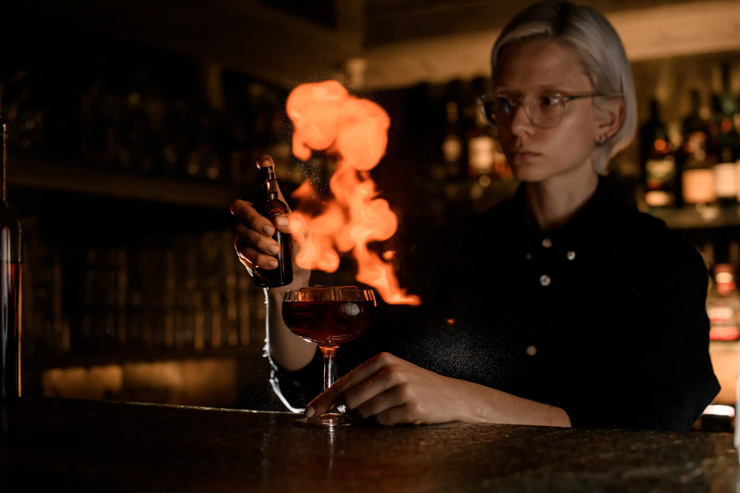 Female bartender in glasses makes fire cocktail with spray and lighted match on bar counter against background of bar showcase