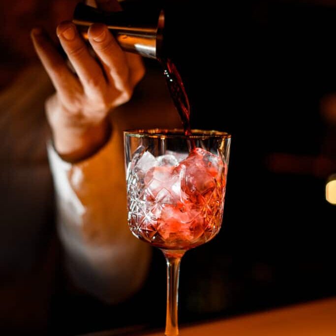 selective focus on crystal glass with ice in which the hand of male bartender pours alcoholic drink from steel jigger