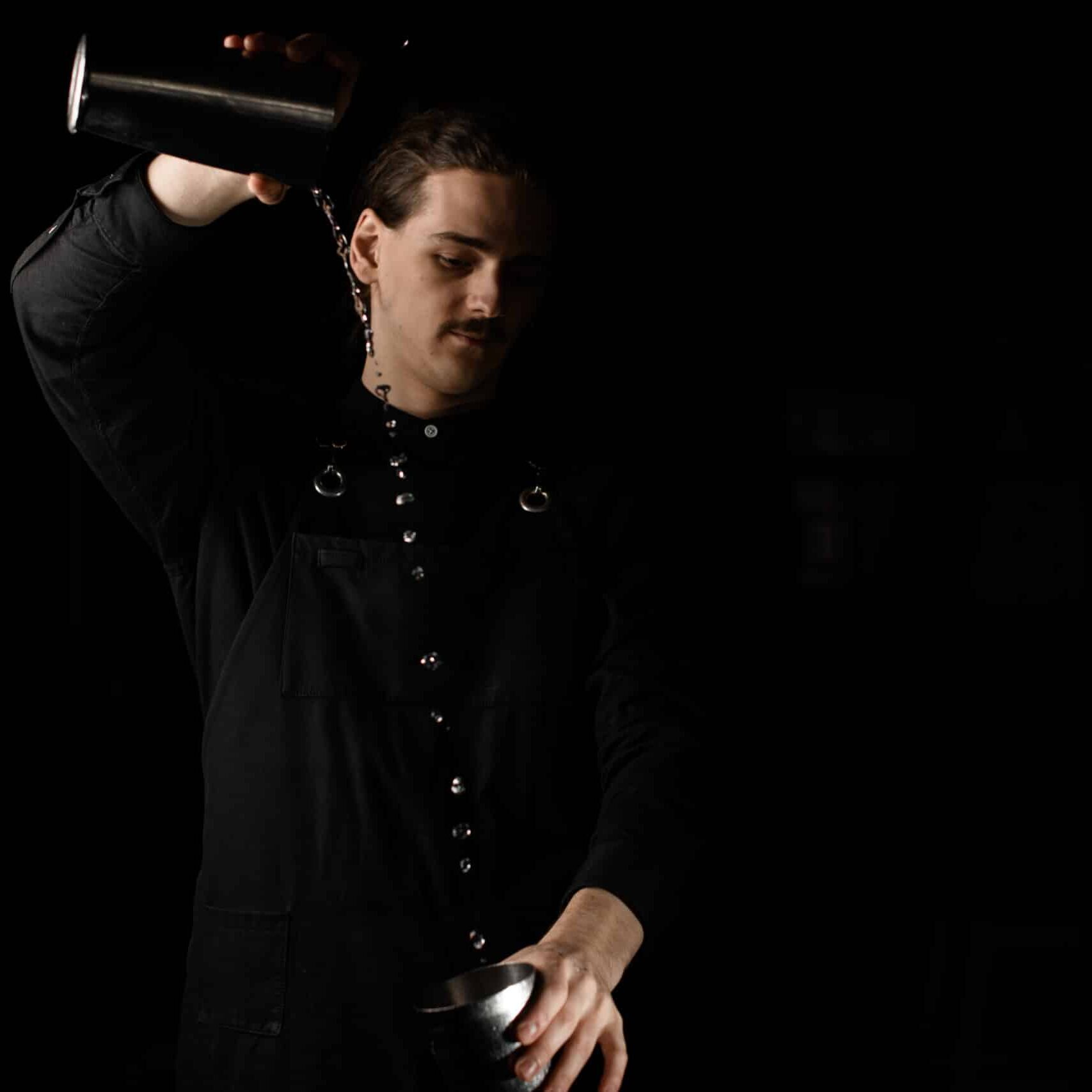 Young smiling bartender pours liquid from one half of a shaker to the other, holding one of them high above his head against a black background